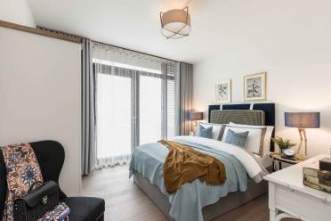A neatly arranged bedroom with a double bed, light blue bedding, two bedside lamps, framed artworks, and a chair with a patterned blanket. The room has floor-to-ceiling windows with sheer curtains.