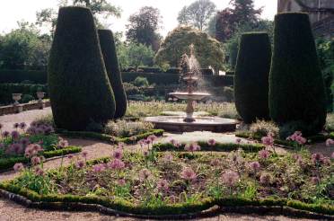 Old Kitchen Garden A formal garden with a central fountain, surrounded by shaped hedges and blooming flowers. Paths and neat garden beds are visible with trees in the background.
