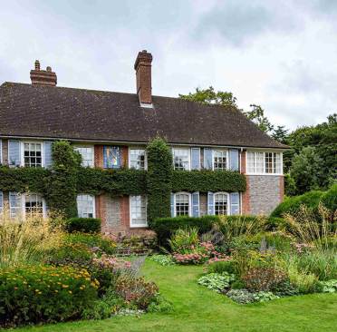 Nuffield Place A two-story brick house with ivy-covered walls and blue shutters is surrounded by a lush garden with various plants and flowers.