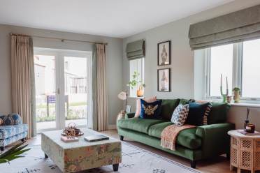 A cozy living room with a green sofa, patterned pillows, a floral ottoman, and a blue chair. Large windows with curtains allow natural light. Wall art and decor complete the space.