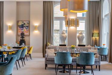 Elegant dining area with blue and yellow chairs, a gray sofa, and round tables set with glasses and flowers. Large windows, artwork, and vases decorate the room. Modern lighting overhead.