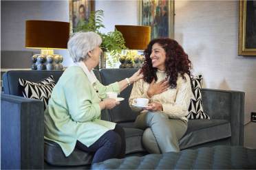 Two women sitting on a sofa holding teacups. One woman, with gray hair, smiles while touching the other, who has long curly hair. They're surrounded by cushions and table lamps.