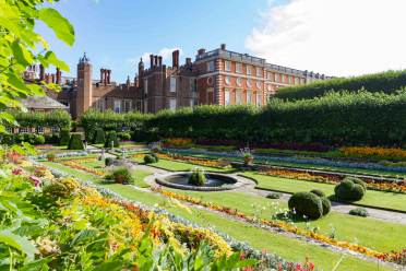 Hampton Court A formal garden with colorful flower beds, trimmed hedges, and a circular fountain in front of a large historic brick building under a partly cloudy sky.