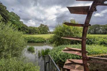 Cobham watermill A large metal water wheel is partially visible on the right, overlooking a river surrounded by green vegetation and trees under a cloudy sky.