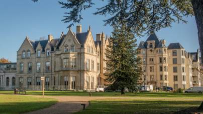 A large historic building with ornate architecture is surrounded by a grassy area, trees, and a clear blue sky.