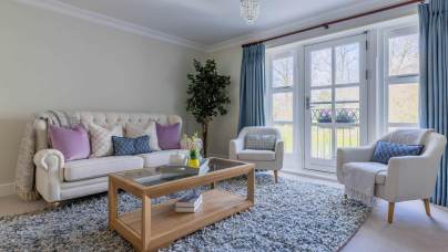 Bright living room with a white sofa and two armchairs, a wooden coffee table, blue curtains, and a large rug. A potted plant is in the corner, and there's natural light from the window.