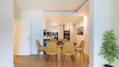 Modern dining area with six mustard-yellow chairs around a wooden table. A kitchen with white cabinets and colorful wall art is visible in the background. A potted plant is on the right.