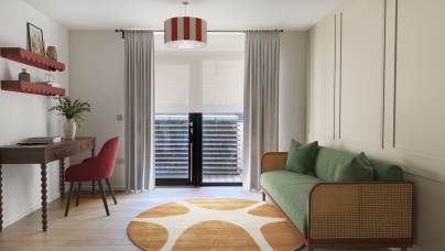 A modern living room with a green sofa, round orange and white rug, a wooden desk with a red chair, and glass doors covered by white curtains leading to a small balcony.