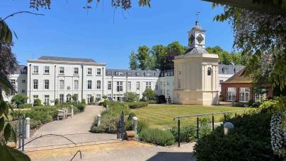 A large white building with many windows and a central clock tower stands behind manicured lawns and pathways under a clear blue sky.