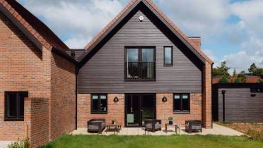 A modern two-story house with dark wood siding, brick walls, patio seating, and green lawn under a partly cloudy sky.