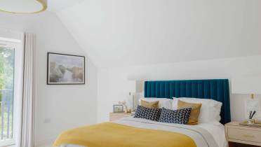 Modern bedroom with a blue upholstered headboard, yellow blanket, decorative pillows, two bedside tables, a large window, and a wall-mounted landscape painting.