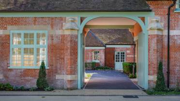 A brick building with an arched entrance leading to a courtyard. It features large windows and a pathway bordered by small plants.