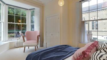 Bright bedroom with a large window, pink chair, and bed with blue and pink bedding. A light fixture hangs from the ceiling, and a building is visible outside through the window.