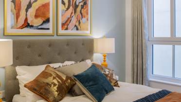 Modern bedroom with a gray upholstered bed, colorful pillows, and two abstract paintings above. Room features large window, white curtains, and bedside tables with lamps.