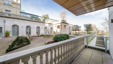 View of a spacious courtyard featuring a stone balustrade, surrounded by a classical building facade with arched doors and a neatly arranged garden.