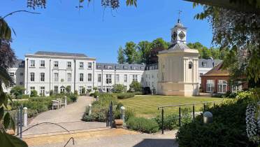 A large white building with many windows and a central clock tower stands behind manicured lawns and pathways under a clear blue sky.