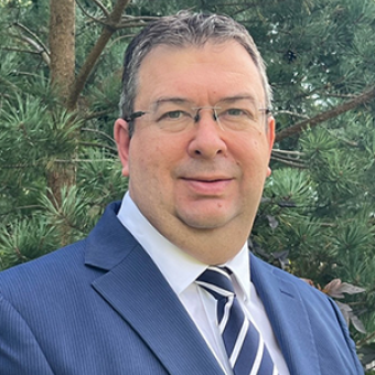 A man in a blue suit, white shirt, and striped tie stands outdoors in front of green pine trees.