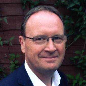 A man with short brown hair and glasses is smiling, wearing a dark jacket and white shirt, standing in front of a wooden fence with green foliage.