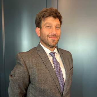 A man with brown hair and a beard wears a grey suit, white shirt, and patterned tie, standing in front of a plain dark wall.