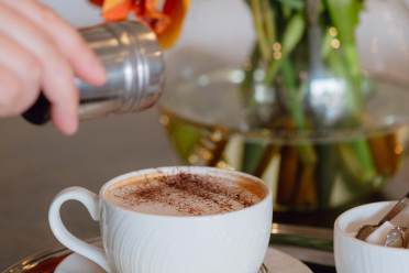 A hand sprinkles cocoa or cinnamon on a cappuccino in a white cup with a saucer, cookie, and spoon. A glass vase with orange flowers is in the background.