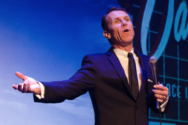 A man in a suit holds a microphone and gestures with one hand while performing on stage in front of a blue backdrop with text.