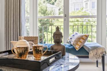 A living room with a marble coffee table featuring a tray with two gold mugs and a teapot, a Buddha statue, a bench with cushions, and large windows showing greenery outside.