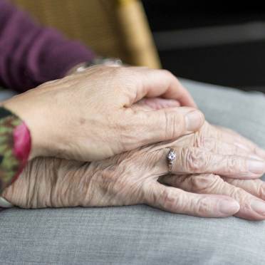 A younger hand gently rests on top of an older hand with a ring, both on a gray fabric surface.
