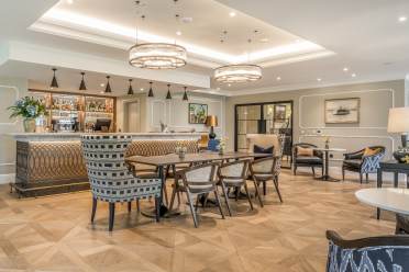 Modern hotel lounge with a bar counter, stylish chairs, a wooden table, and contemporary lighting fixtures. The space is decorated with art, plants, and a neutral color palette.