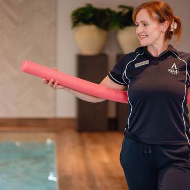 A woman in a uniform holds a pink foam pool noodle next to an indoor pool, smiling and looking to the side.
