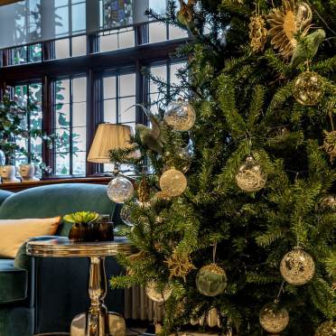 A decorated Christmas tree with gold and silver ornaments is next to a round side table and a green sofa in a room with large windows.