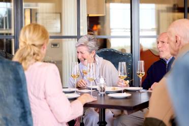 Four elderly people sit at a restaurant table, engaged in conversation, with wine glasses in front of them. The setting is bright and relaxed.