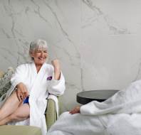 An older woman in a white robe sits on a sofa, smiling and talking with another person who is also in a robe, in a modern, marble-walled room.