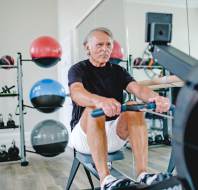 An older man in athletic clothing uses a rowing machine in a gym with kettlebells and medicine balls on racks in the background.
