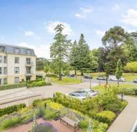 View of a modern apartment building with balconies, landscaped gardens, parked cars, and a background of large trees under a partly cloudy sky.