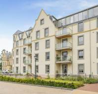 Modern multi-story apartment building with cream facade, balconies, and large windows. A small garden is in front, and there's a parked car on a driveway to the left.