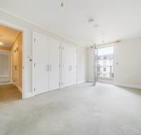 Empty, carpeted bedroom with white walls, three built-in closets, and a door open to a balcony, letting in natural light. Hallway visible on the left.