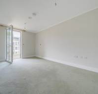 Empty, unfurnished room with light-colored walls and carpet, built-in wardrobe on the left, and an open glass door leading to a balcony with a view of buildings outside.