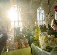 People browse various handmade goods at indoor market stalls, with sunlight streaming through large arched windows, illuminating the room.