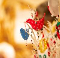 Fabric birds in red and blue with patterns and beads hang on a white decorative tree, with a blurred golden background.