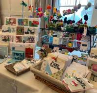 A display table with handmade greeting cards, pop-up cards, and crochet decorations at a craft fair, with natural light from nearby windows.
