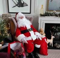 A person in a Santa Claus costume sits in an armchair next to a decorated Christmas tree and fireplace with holiday decorations.