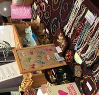 A display table features assorted handmade jewelry, including beaded necklaces, earrings, and bracelets, with signs and information sheets visible in the background.