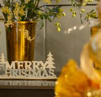 A glittery "Merry Christmas" sign sits on a surface next to a gold vase with festive greenery; blurred gold decorations appear in the foreground.