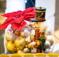 A wooden nutcracker figurine stands in front of a glass bowl filled with gold and silver ornaments, with a large red poinsettia flower behind it.