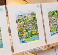 Three colorful, framed prints of illustrated landscapes are displayed upright on a wooden shelf in an indoor setting.