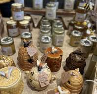 Beehive-shaped ceramic honey pots and jars of honey are displayed on a table with handwritten labels, in front of a chalkboard sign.