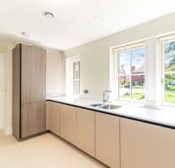 Modern kitchen with light-colored cabinets, a built-in tall cupboard, stainless steel sink, large windows, and view of a green outdoor area.
