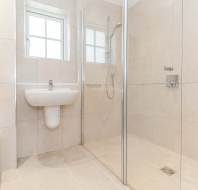 Modern bathroom with beige tiles, a wall-mounted sink, shower area with glass partition, and toilet on the left. A window lets in natural light.