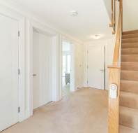 A bright hallway with beige carpet, three white doors, a staircase with a wooden handrail, and a light fixture on the ceiling.