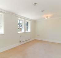 Empty, light-filled room with cream walls, beige carpet, three windows, a radiator, and a ceiling light fixture.